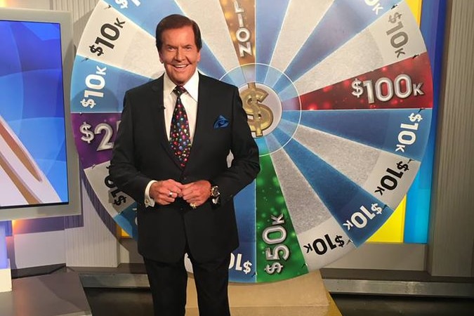 A smiling man in a suit stands into front of a sparkling wheel displaying numbers relating to cash amounts to win.