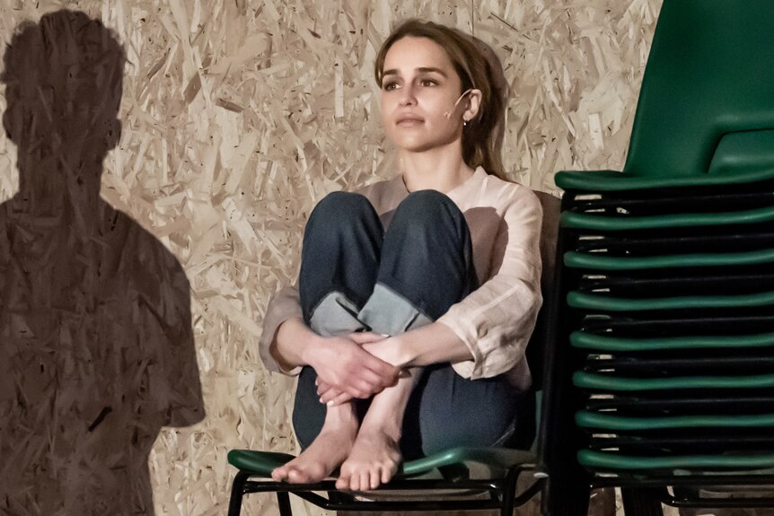 A young, smartly dressed blonde woman sits in a green chair, her knees up against her chest, looking morose