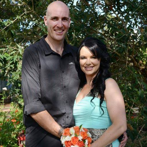 A tall man and a woman stand together closely under a tree.