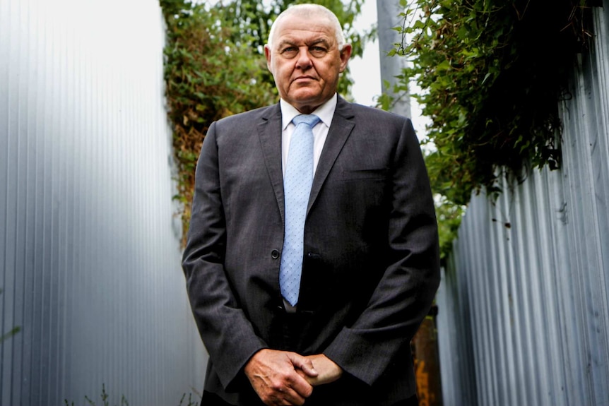 Ron Iddles, wearing a suit and tie, stands in a laneway and looks directly at the camera.