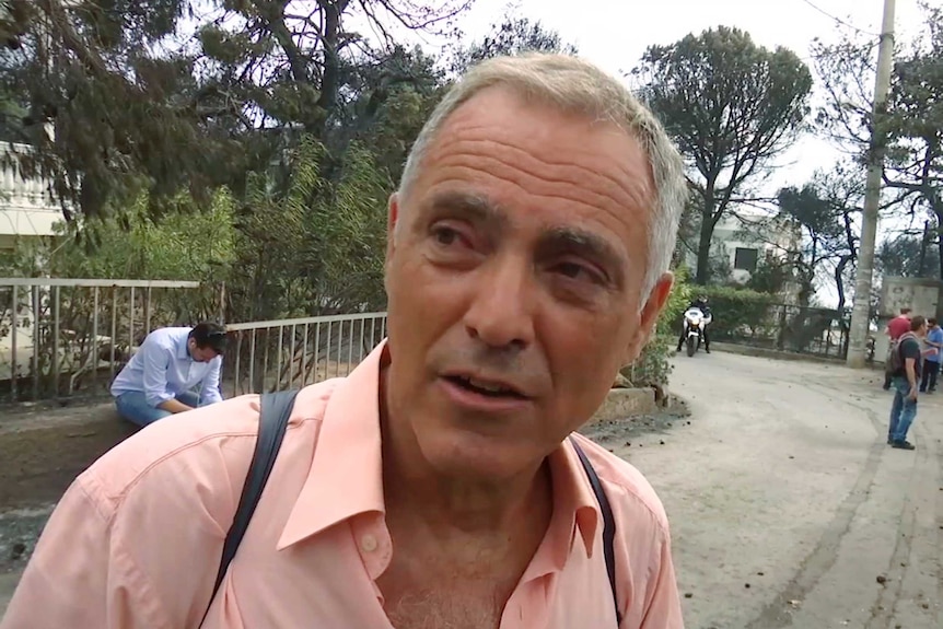 Nikos Stavrinidis, wearing a pink shirt, looks to the left of the screen.