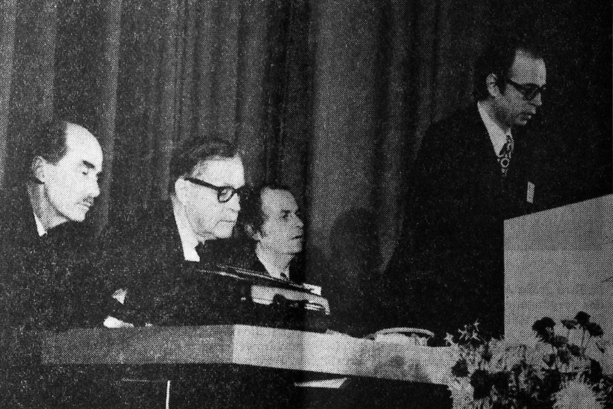 A group of men in suits sit behind a mand standing at a lectern. The photo is in black and white.