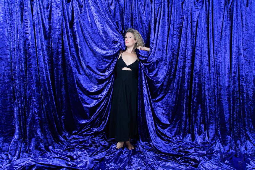 Woman with curly blonde hair and blue eyes stands in a blue velvet curtain animated with movement
