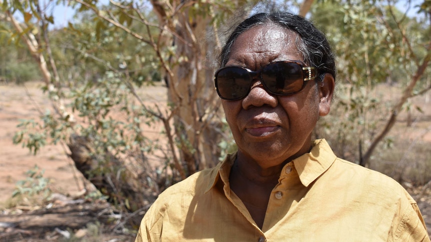 Margie Lynch is wearing a yellow shirt and sunglasses and looks with a serious expression at the camera.