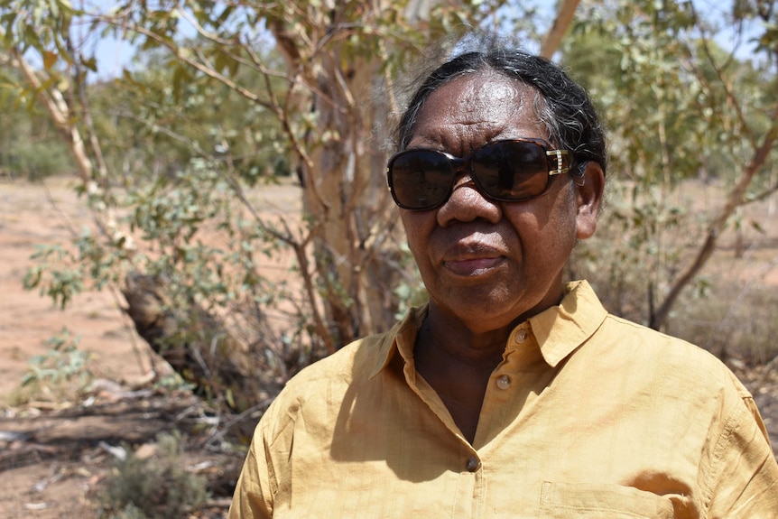 Margie Lynch is wearing a yellow shirt and sunglasses and looks with a serious expression at the camera.