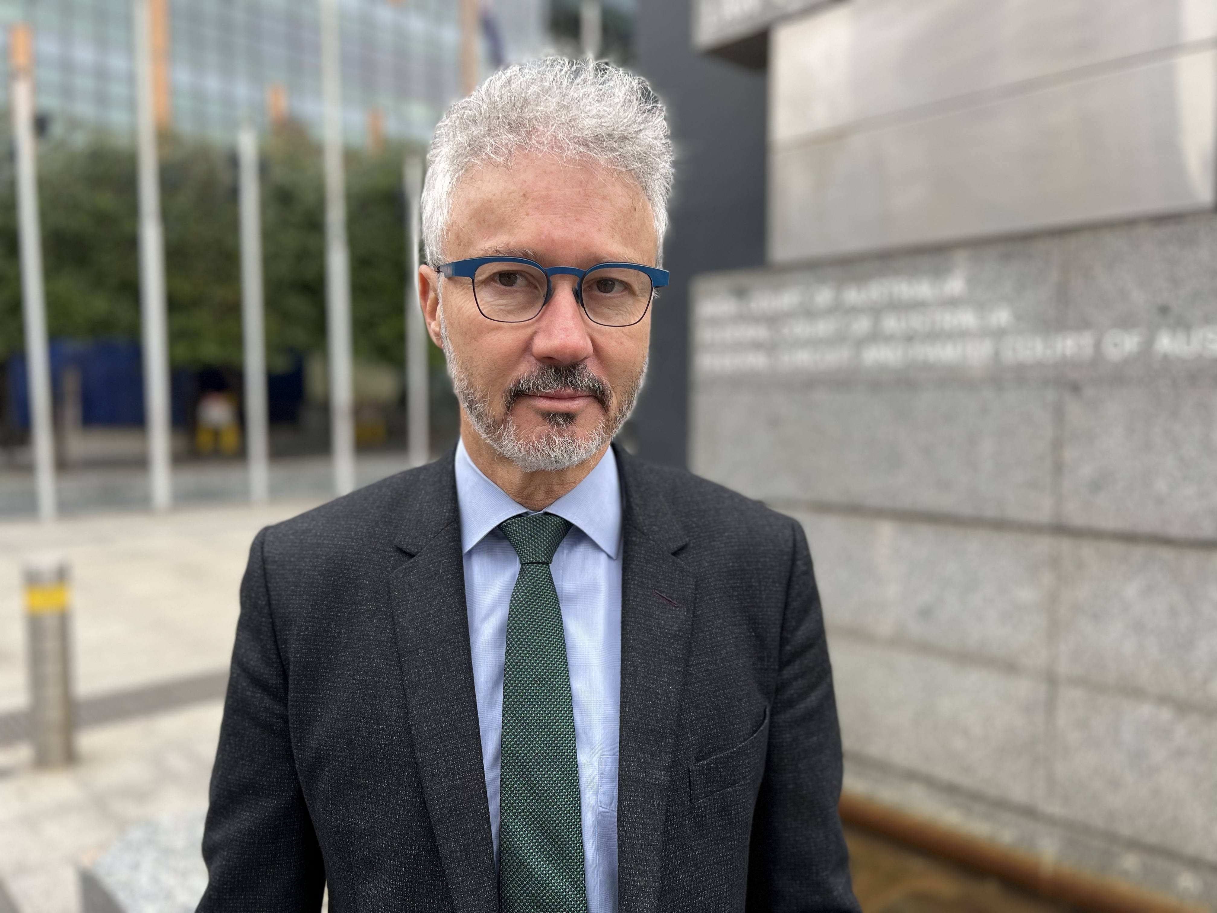 A man in a suit with short gray hair and glasses stands outside the court