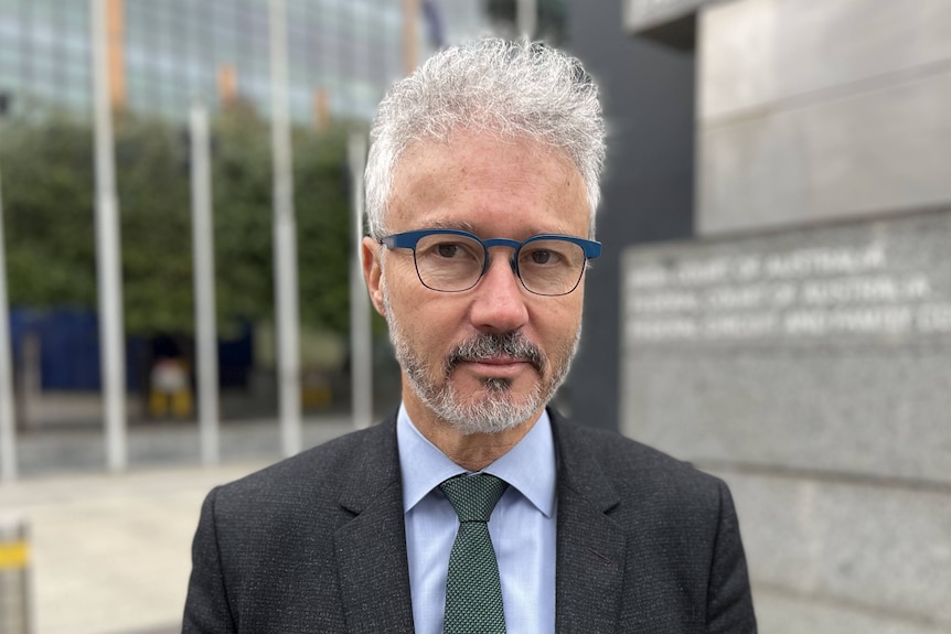 A man in a suit with short gray hair and glasses stands outside the court