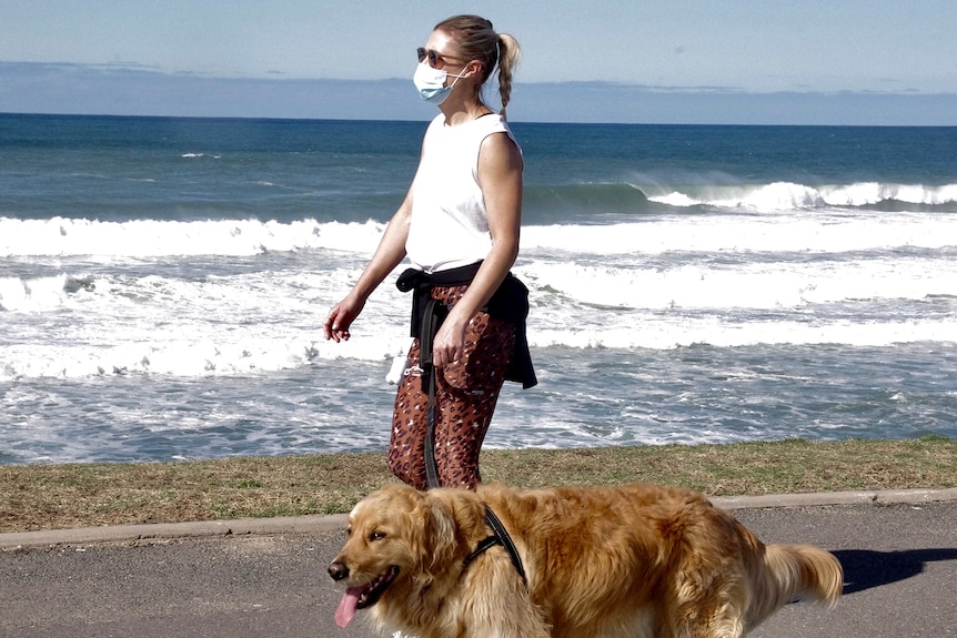 A woman wearing a face mask walks a god along a beach.