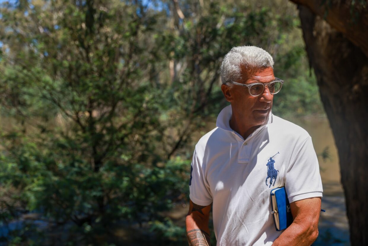 An Indigenous man with white hair, standing on a river bank.