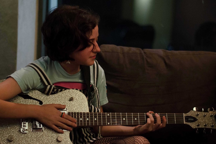 A young woman sits on a couch playing a sparkly guitar