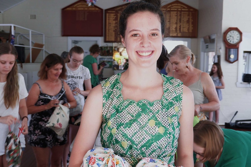 Lily Harrison looks to the camera as she holds to floral bags in her hand. She smiles, and is wearing a green top.