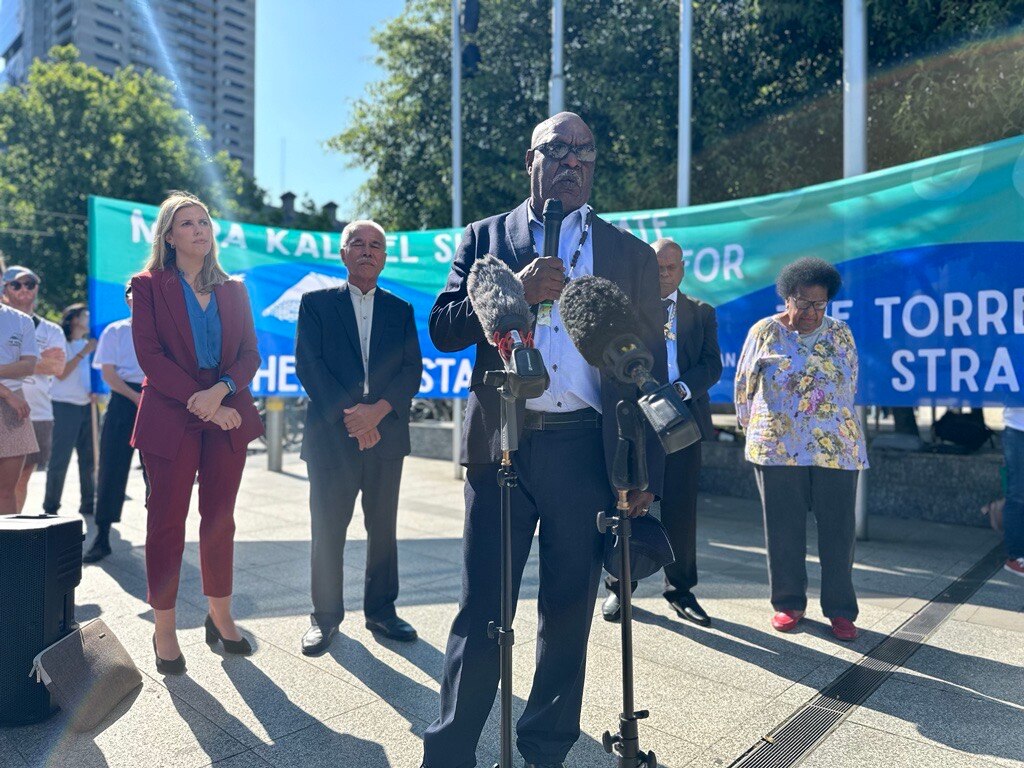  man in a suit stands in front of microphones in front of the torres strait flag banner
