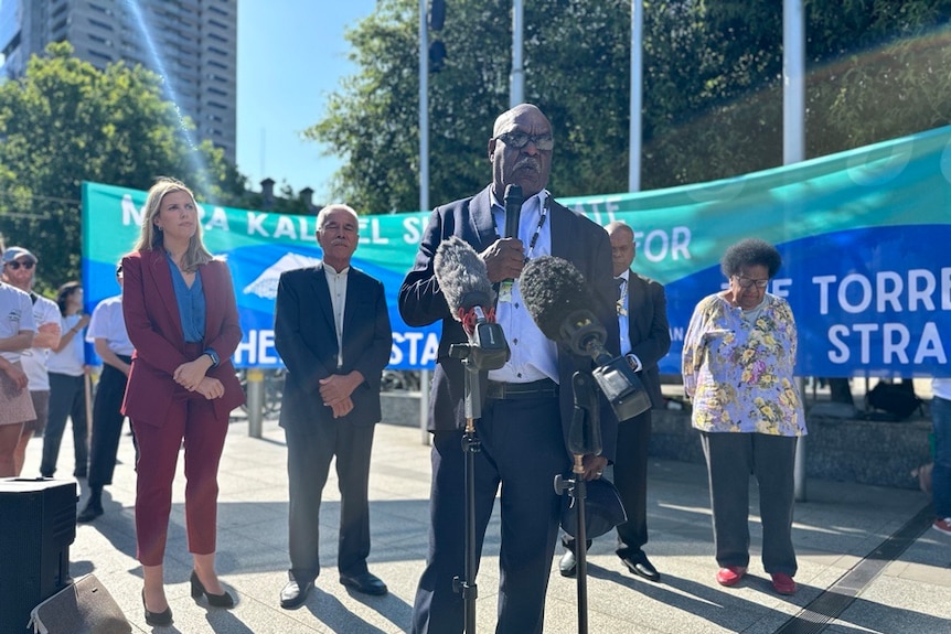  man in a suit stands in front of microphones in front of the torres strait flag banner