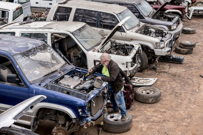 A man with a cigarette in his mouth stands on a wheel while working on an old car engine at the auto wreckers.