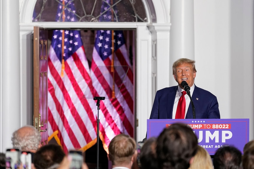 Donald Trump grins while addressing a crowd from behind a lectern with US flags