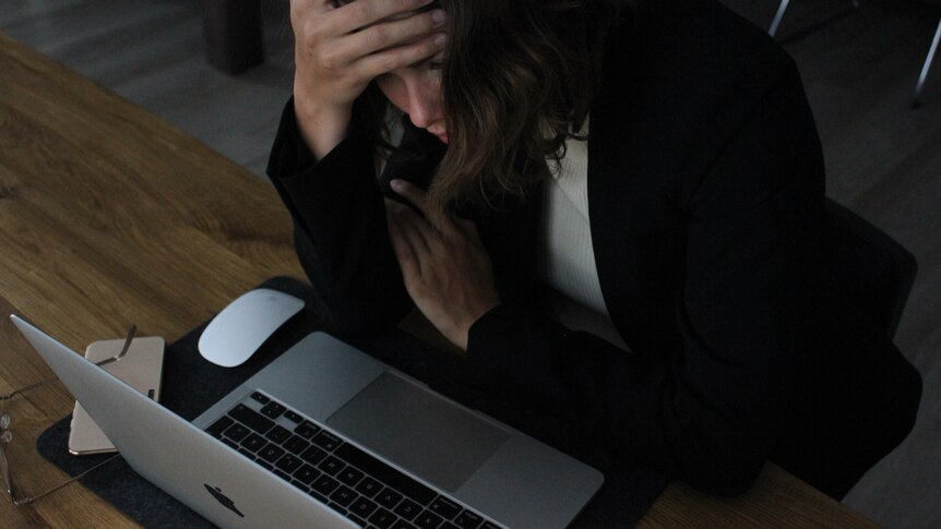A woman with her hand on her forehead looking at an apple laptop, appearing stressed. 