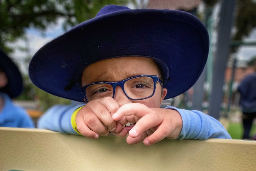 A picture of a young boy with a hat and glasses looking into the camera from behind a piece of playground equipment.