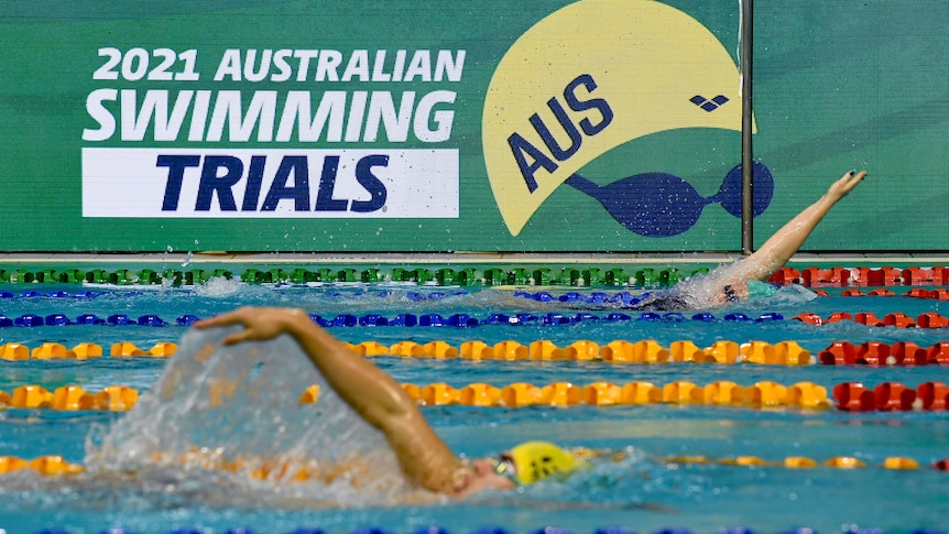 Swimmers go through their paces in the pool while in the background lies a big sign "2021 Australian Swimming Trials".