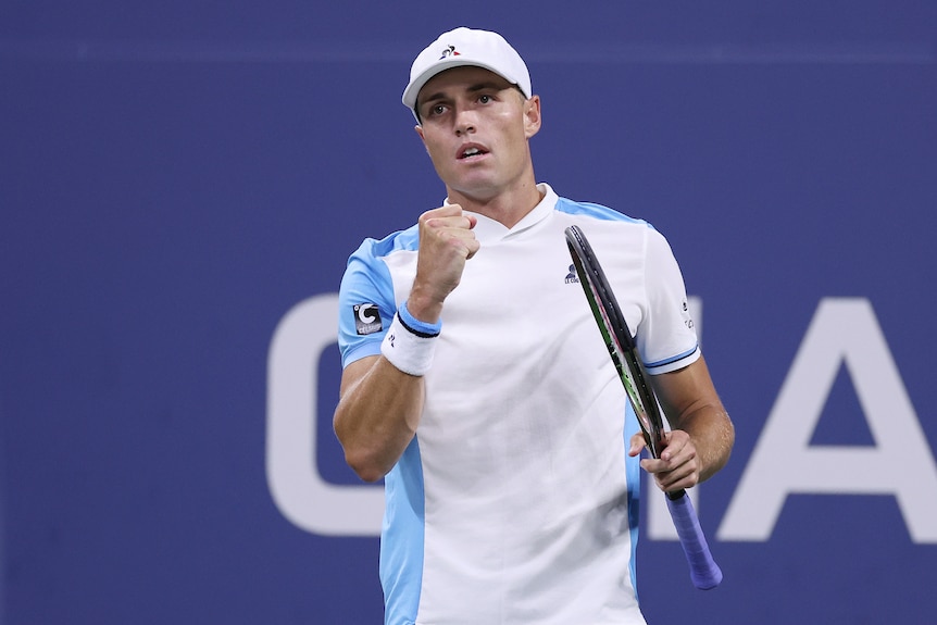 A male tennis player in a white shirt and white cap, pumps his fist after winning a point.