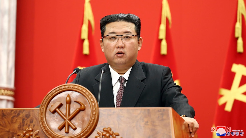 In a dark suit with white shirt and red tie, Kim Jong Un gives speech at brown lecturn in front of red and yellow lecturn.