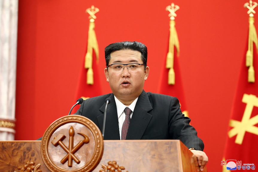 Indossando un abito scuro con una camicia bianca e una cravatta rossa, Kim Jong-un sta tenendo un discorso in una conferenza marrone davanti a una conferenza rossa e gialla.