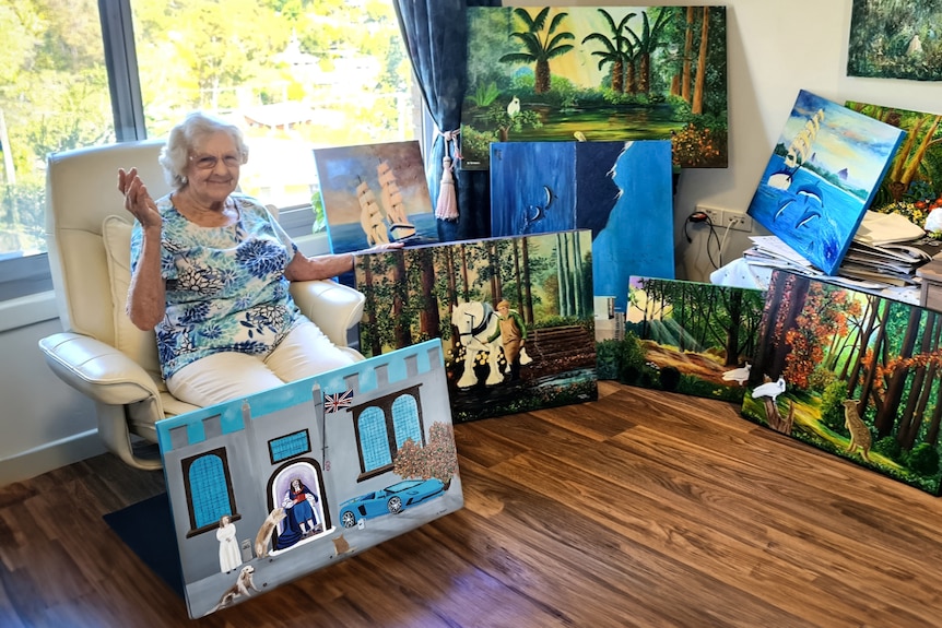 An elderly woman sits in a sunny room with several of her brightly coloured paintings.