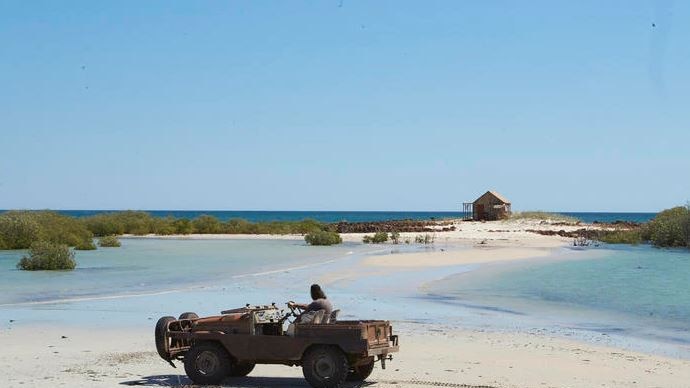 A man in a roofless jeep on the beach looks across a shallow sandbar to a timber shack surrounded by blue water and mangroves