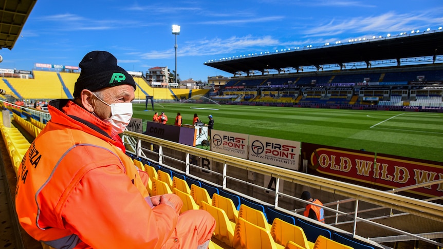 A steward sits in a football stadium with an orange coat on and a face mask