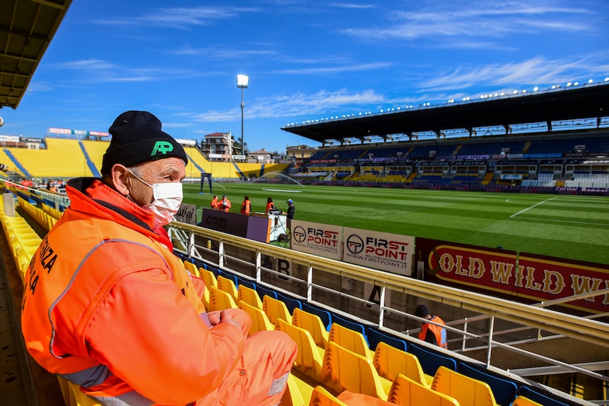 A steward sits in a football stadium with an orange coat on and a face mask