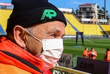 A steward sits in a football stadium with an orange coat on and a face mask
