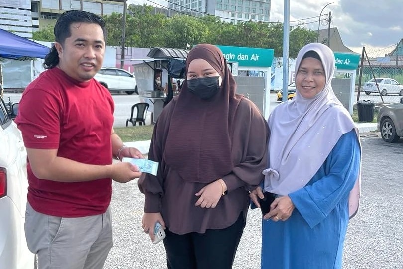 A man in red T-shirt stands holding a bank note next to two Muslim women in street