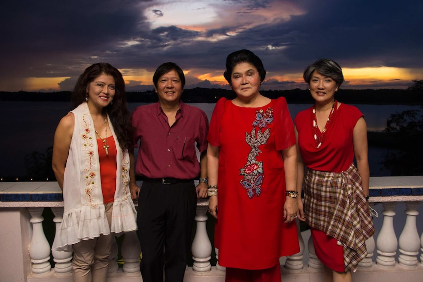 Four members of the Marcos family stand in in front of a white, neoclassical balustrade in front of a moody sky.