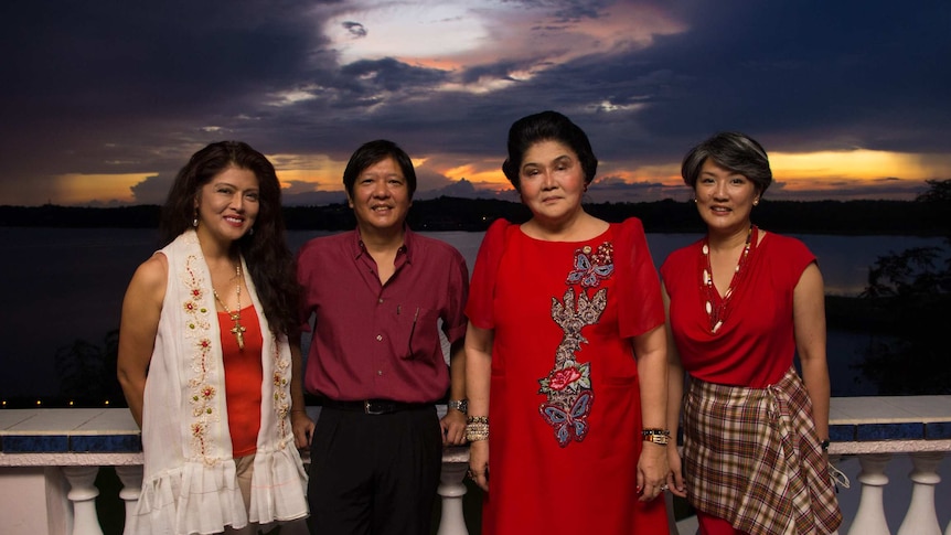 Four members of the Marcos family stand in in front of a white, neoclassical balustrade in front of a moody sky.