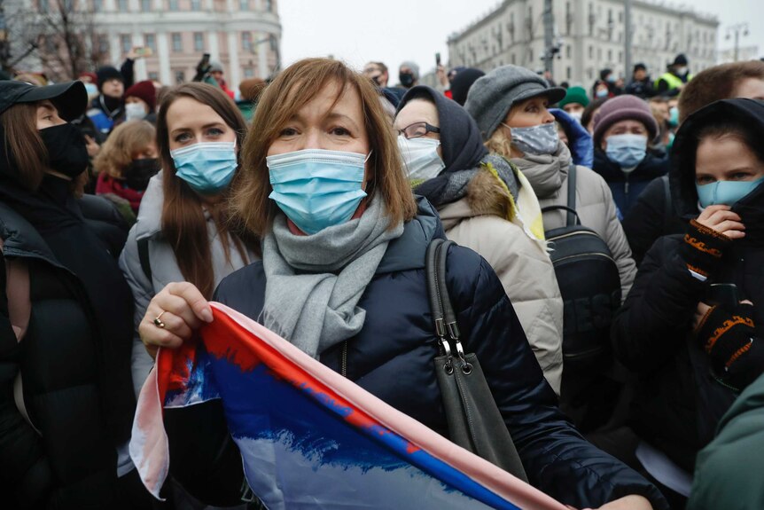 Women wearing face masks stand in a crowd, some carrying a flag