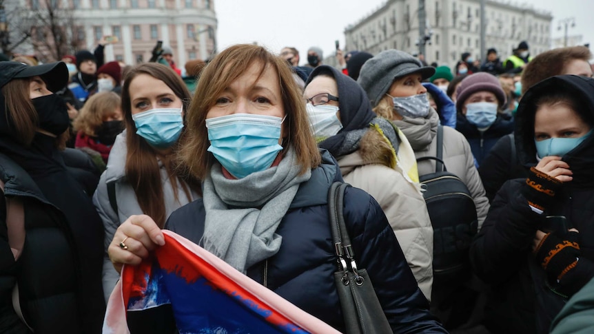 Women wearing face masks stand in a crowd, some carrying a flag