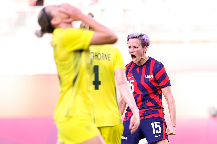 A soccer player celebrates while a defender hold her head in her hand in the foreground.