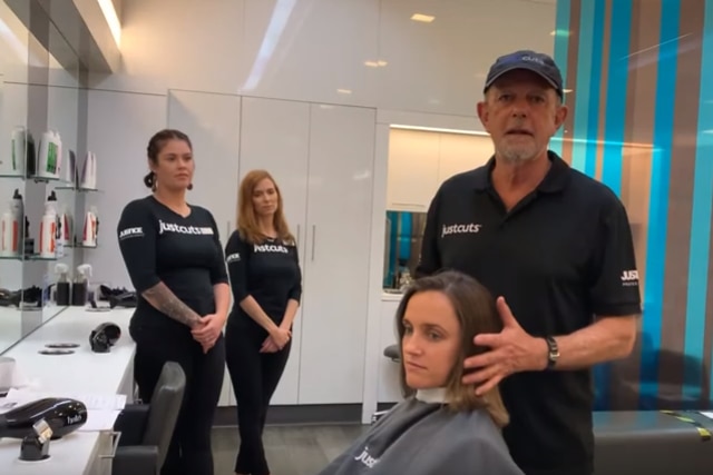 A man in a Just Cuts t-shirt touching a client's hair, with two women in background