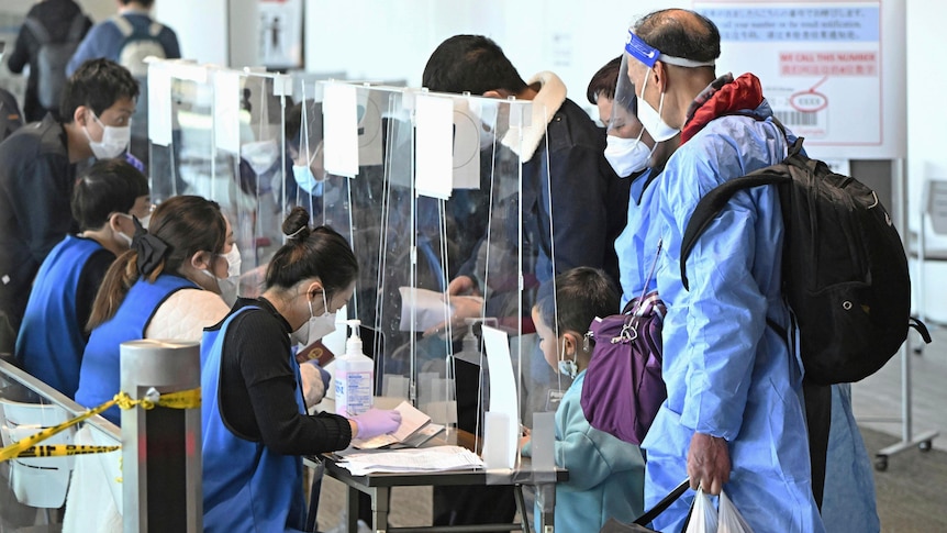 Passengers in PPE and face masks show documents to clerks seated behind clear protective barriers.