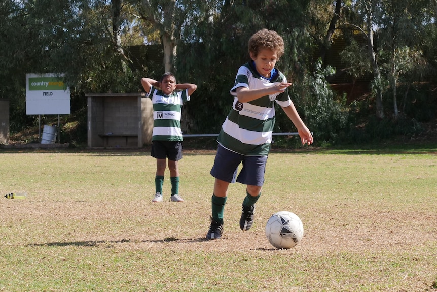 Un garçon, la langue tirée, pointe sa botte vers le ballon de football tandis qu'un autre enfant, les mains sur la tête, regarde.