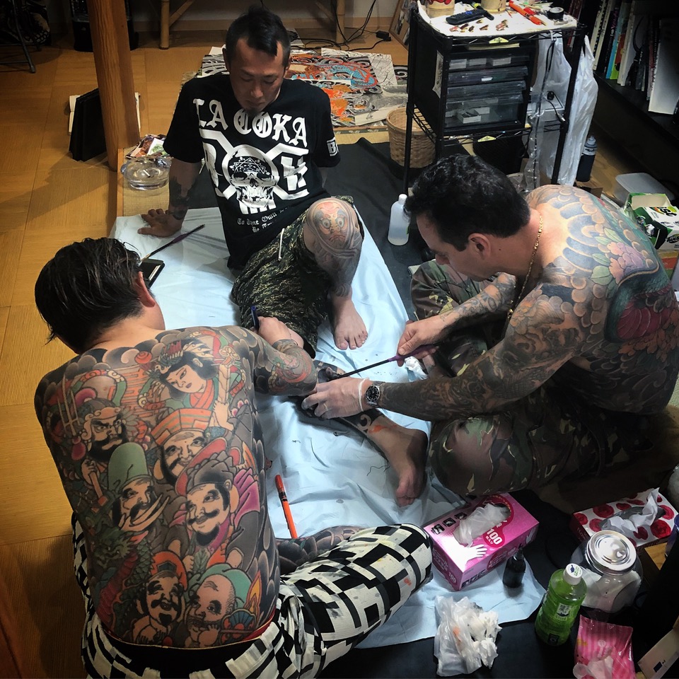Two topless men with lots of traditional tattoos carry out the tebori method of tattooing on a japanese man