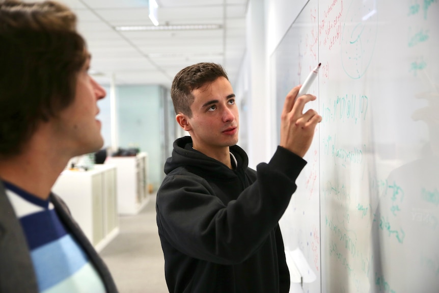 A yong man points to a whiteboard with a whiteboard marker while another man looks on.