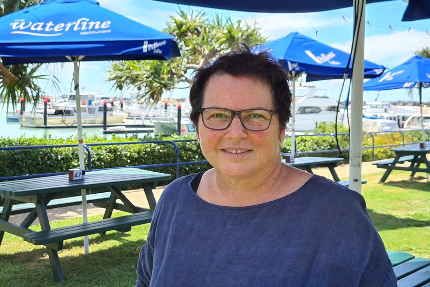 A woman wearing a blue shirt smiles. There is empty tables in the background, with a harbour behind them