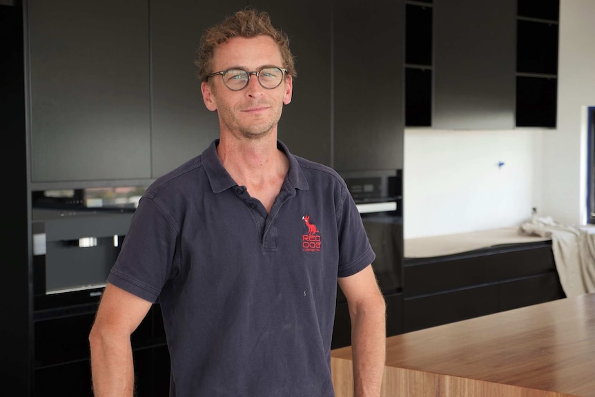 Man with spectacles wearing a navy blue polo shirt standing in new home's kitchen.