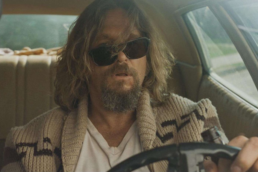 Jeff Bridges sits behind the wheel of a car. His hair is across his face, his sunglasses are askew and his mouth slightly ajar.