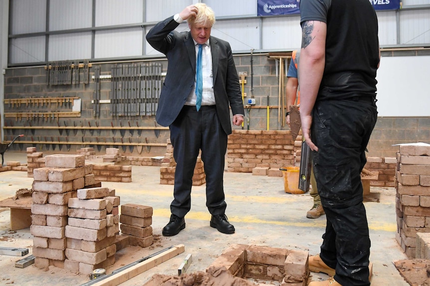 Boris Johnson, wearing a suit and tie scratches his head and looks at bricks on the ground.