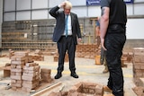 Boris Johnson, wearing a suit and tie scratches his head and looks at bricks on the ground.