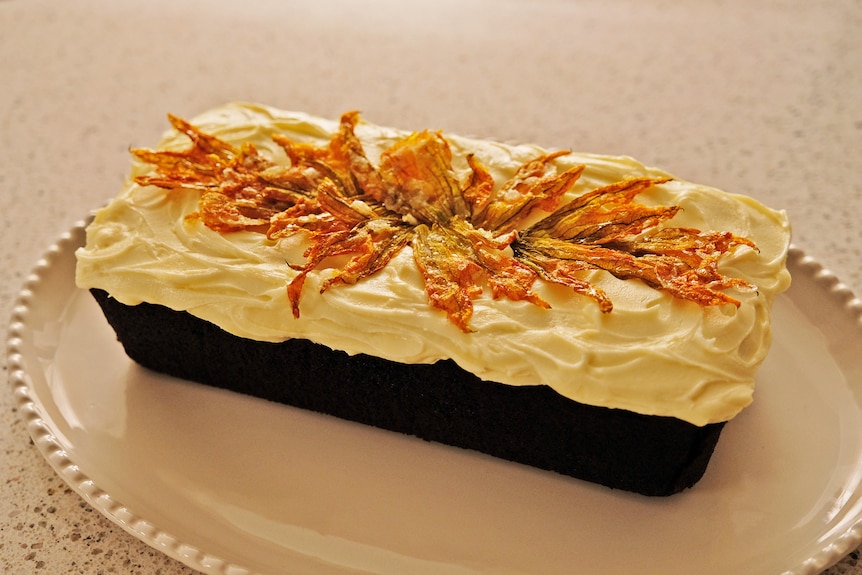 A loaf of chocolate zucchini cake with cream cheese icing and candied zucchini flowers served on a white dish.