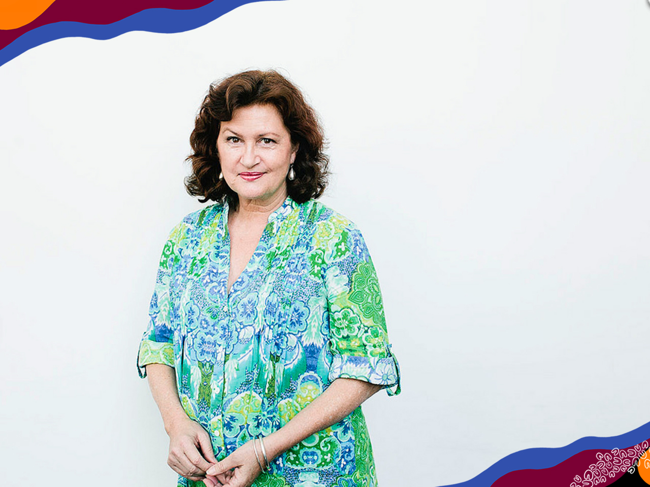 A woman with brown curly bob in lime green and blue patterned shirt stands in front of white wall.