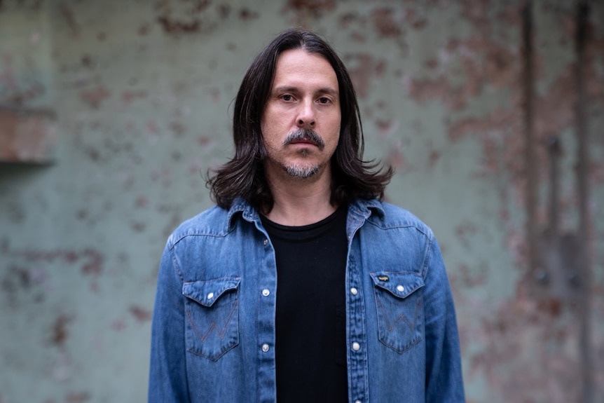 Man with dark hair sitting just above the shoulders looks serious down the barrel. He wears a denim jacket.