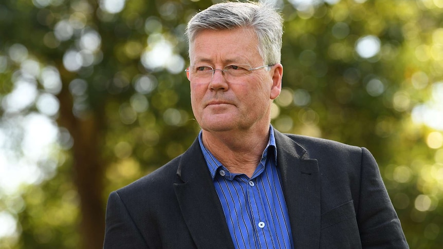 A bespectacled man with salt-and-pepper hair wearing a blue shirt and dark jacket is photographed in front of blurred foliage